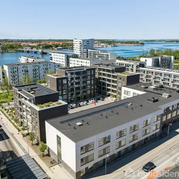 Bygninger ligger ved en sø, omgivet af flere moderne lejlighedskomplekser i forskellige højder. Gader løber mellem bygningerne med biler parkeret langs vejene. Længere væk ses en bysilhuet.