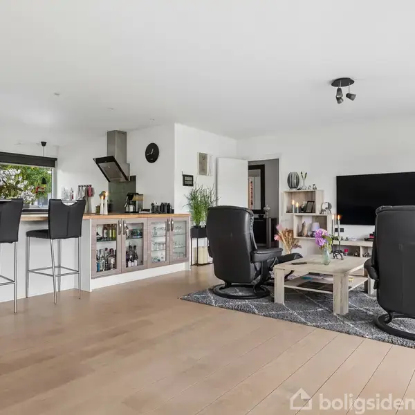 Stue med åbent køkkenalrum. To sorte lænestole vender mod en stor fladskærm. Køkkenø med tre barstole til venstre; bord med blomster og sofaer i midten.