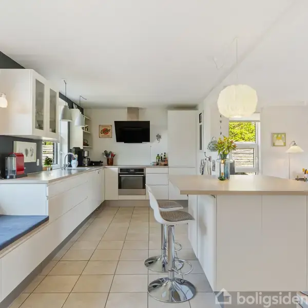 Køkken med hvide skabe og moderne apparater, barstole omkring en køkkenø; lyst og rummeligt med store vinduer, sidelæns siddeplads og udsigt til en stue med sofa og kunst på væggene.