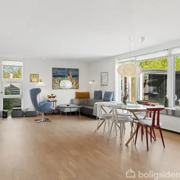 En lys stue med trægulv indeholder spisebord med stole, en grå sofa ved hjørnet og en blå lænestol. Store vinduer slipper naturligt lys ind fra haven udenfor.