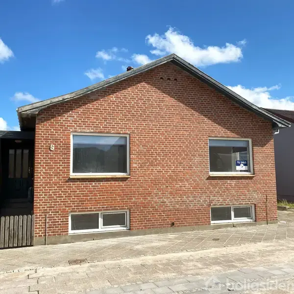 En rød murstensbygning med to større vinduer foran. Skilt i det højre vindue viser "Til salg". Bygningen står i et boligkvarter under en klar blå himmel.