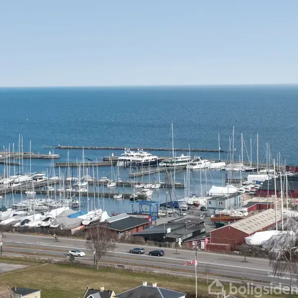 Både ligger fortøjet i en lystbådehavn ved kysten. I baggrunden ses det åbne hav, mens veje og bygninger omgiver havnen i forgrunden.