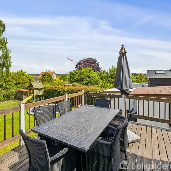 En udendørs terrasse med et spisebord omgivet af seks stole og en lukket parasol. I baggrunden ses en velplejet have med buske, græsplæne og en flagstang.