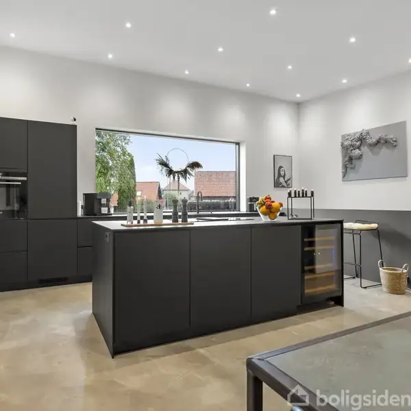 Moderne køkken med mørkegrå skabe og central ø med barstole. Store vinduer giver udsigt til træer og røde tage. Rum er lyst op med loftsindbyggede spotlys.
