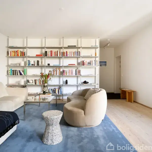 Reol fyldt med bøger og dekorationer i moderne stue. Beige sofa og stol står på blåt tæppe. En lav bordplade rummer vaser med blomster. Lys trægulv, hvid væg med indrammet billede.