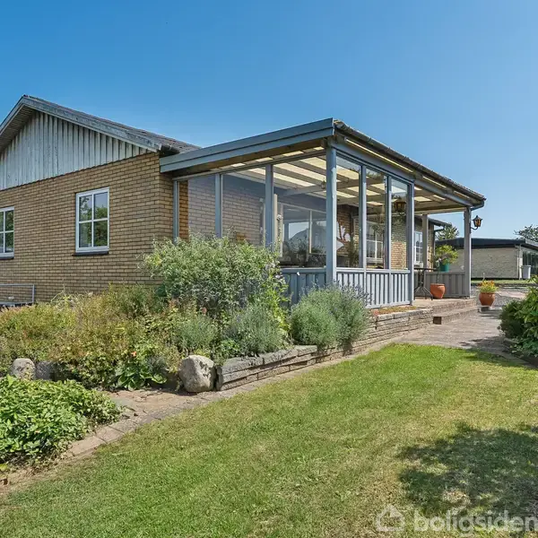 Hus med murstensfacade har en glasinddækket terrasse. Det ligger i en have med græsplæne og buske, under en klar blå himmel.