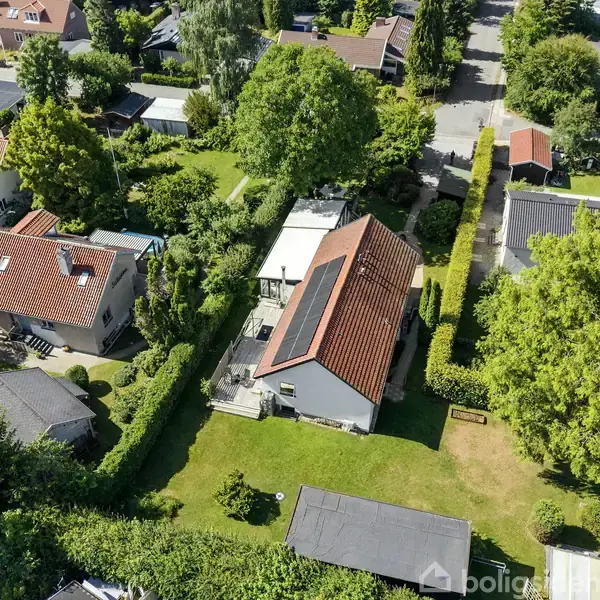 Et hus med rødt tegltag og solpaneler står i en grøn have. Omgivet af hække og træer ses flere huse og veje i det tætbefolkede nabolag.