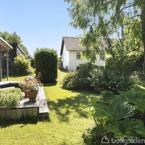 Træhus med pergola og liggestol står i en frodig have med græsplæne, store buske og træer. I baggrunden ses et hvidt hus med tegltag.