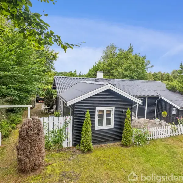 Sort træhus med hvid terrasse og hegn. Det står midt i en grøn have omgivet af høje træer og buske. Himlen er klar blå.