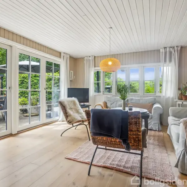 Stue med hvide lofter og trægulv, fyldt med møbler som sofaer og stole, og en rund bordlampe. Store vinduer åbner ud til en terrasse med udsigt til grønne omgivelser.