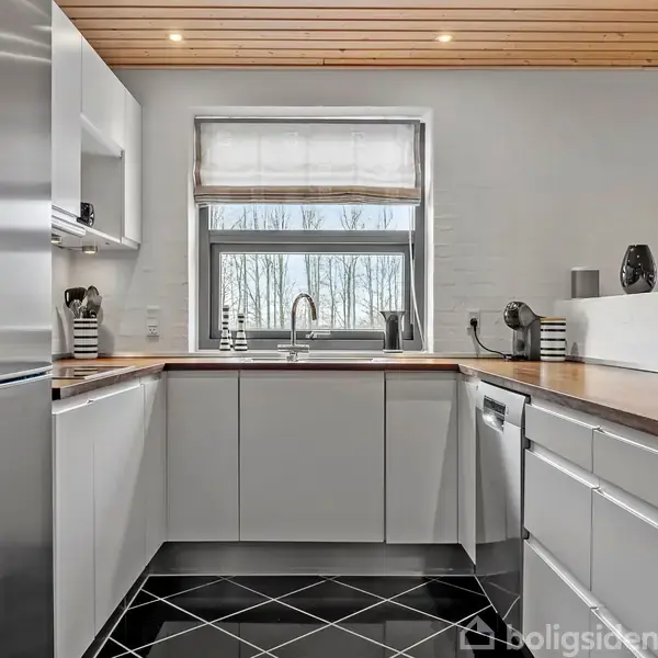 Et moderne køkken med hvide skabe, der står stille med stålgreb. I centrum er en vask monteret under et stort vindue. Sort flisegulv giver kontrast til lyse trælofter.