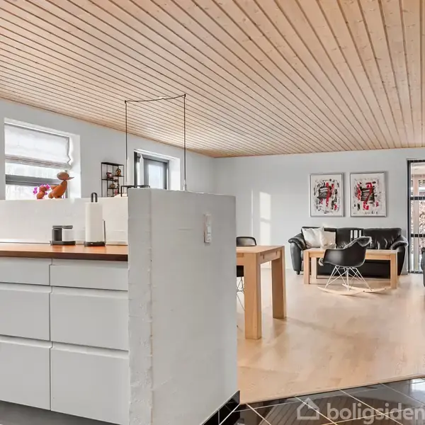 Et moderne køkken med hvide skabe åbner op til en lys stue med trægulv og -loft, indeholdende et træbord, sorte lædersofaer og dekorative billeder på væggen.