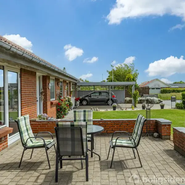 Terrassemøbler står på en brolagt terrasse ved siden af et murstenshus. I baggrunden er der en græsplæne og en parkeret bil ved en carport. Klar blå himmel med skyer.