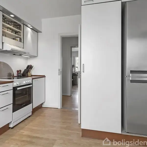 Et moderne køkken med hvide skabe og stålgreb. Komfur og ovn ved siden af åbne skabsdøre med glas. Trælaminatgulv og døråbning til en lys stue i baggrunden.