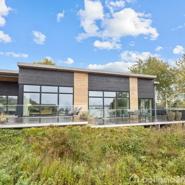 Moderne hus med store glaspartier står på hævet terrasse. Det er omgivet af frodig vegetation. Himmel med spredte skyer skaber en lys og naturlig atmosfære.