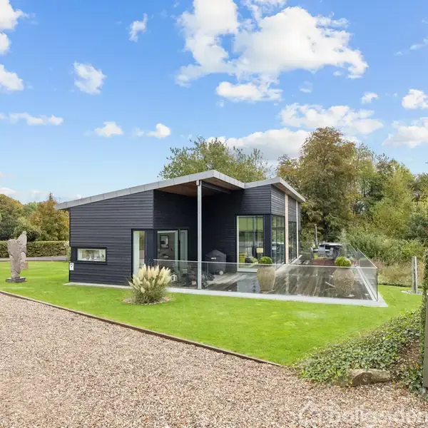 En rund, udendørs jacuzzi står på en terrasse med glasrækværk. I baggrunden ses en udsigt over en sø, grønne bakker og et hus. Himlen er delvist overskyet.