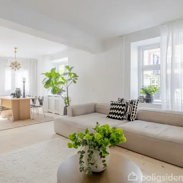 En lys stue med en beige sofa og sorte/hvide puder. En grøn plante står ved sofabordet. I baggrunden ses et spisebord med seks stole under en dekorativ lampe.