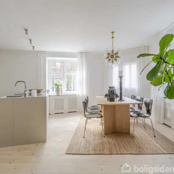 Et moderne køkken med en ø i midten har et spisebord til seks personer. Stort vindue bagved giver naturligt lys, og indretningen er lys med grønne planter.