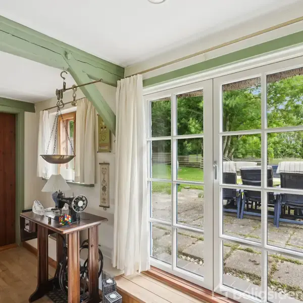 En antik symaskine står på et træbord. Bagved er en stor glasdør med udsigt til en terrasse med havemøbler og en grøn have. Rummet har træbjælker og lyse gardiner.