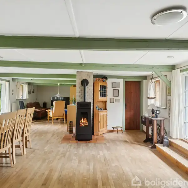 En stue med brændeovn i midten afgiver hyggelig varme. Til venstre står et langbord med stole. Store vinduer til højre giver udsigt til haven.