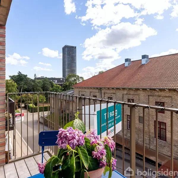 En balkon med farverige blomster i en potte på et blåmalet bord. Udsigten viser en gade, bygninger og en høj bygning i baggrunden under en blå himmel.