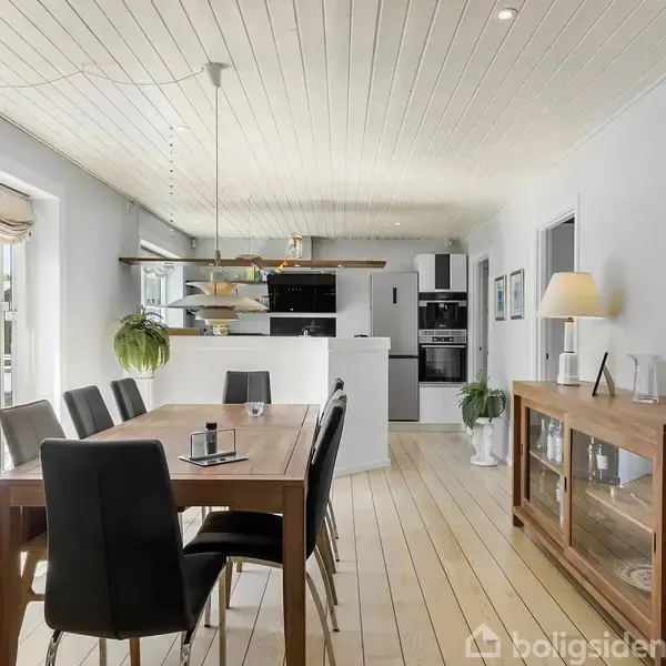 Spisebord med sorte stole står i et lyst køkken-alrum. Lamper hænger over bordet, mens hvidevarer og dekorative elementer som planter og lamper pryder rummet. Store vinduer giver naturligt lys.