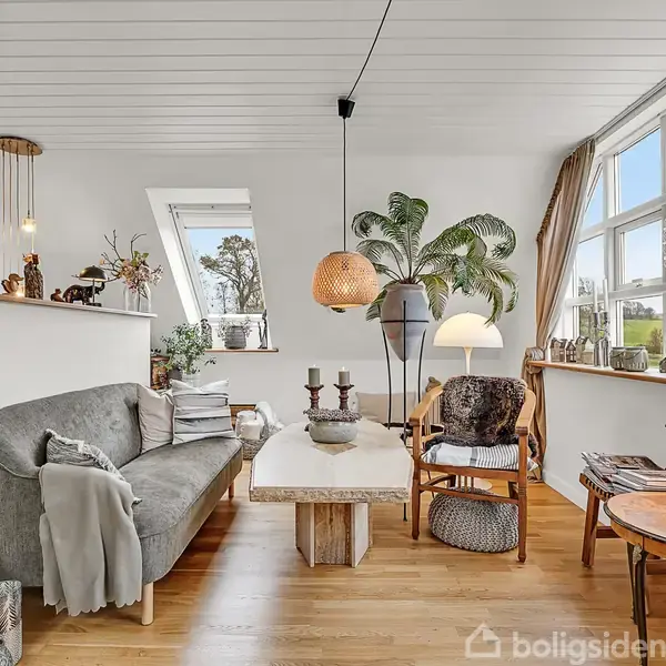 Sofa står i en stilfuld stue, omgivet af planter og moderne lamper, der skaber en lys og hyggelig atmosfære. Store vinduer giver udsigt til træer udenfor.