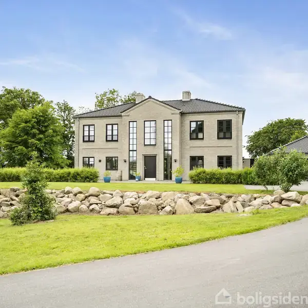 Et stort, moderne hus står roligt omgivet af en velplejet have og grønne træer. Huset har rene linjer og store vinduer. Der er en stenmur og en grusvej foran.