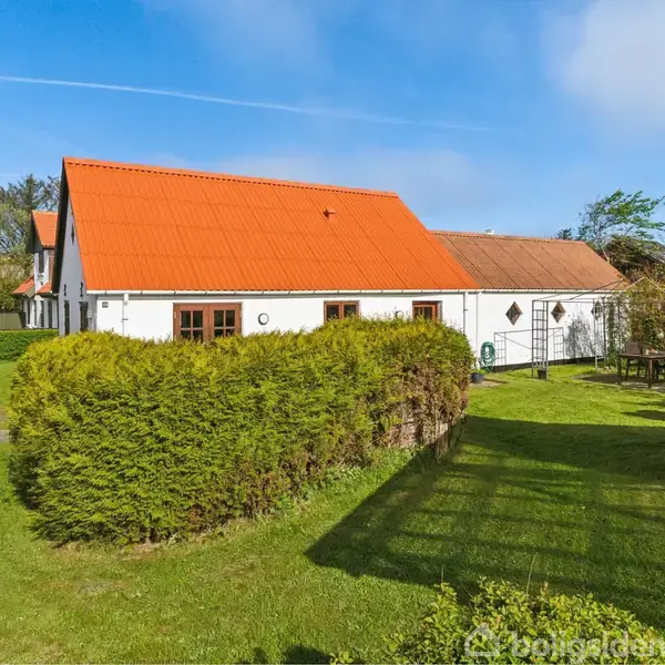 Hus med rødt tag står stille omgivet af grønne hække og græsplæne i et solrigt villakvarter.