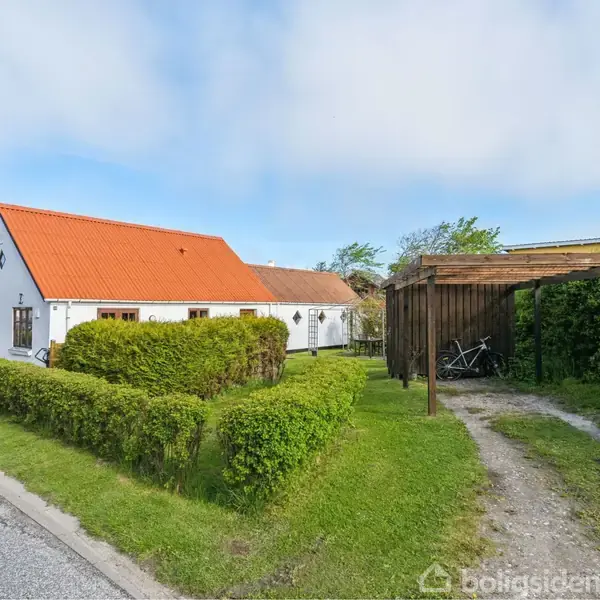 Et hvidt hus med rødt tag står på en stille gade, omkranset af en grøn hæk og en carport med cykler ved siden af.