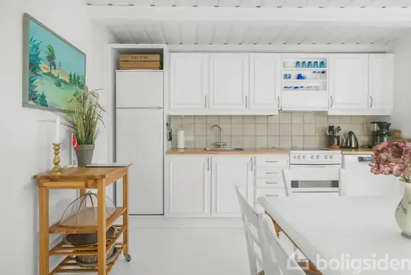 Hvidt køkken med skabe og komfur. Plante på et træbord til venstre. Billedkunst på væggen. Vase med blomster på spisebordet i centrum.