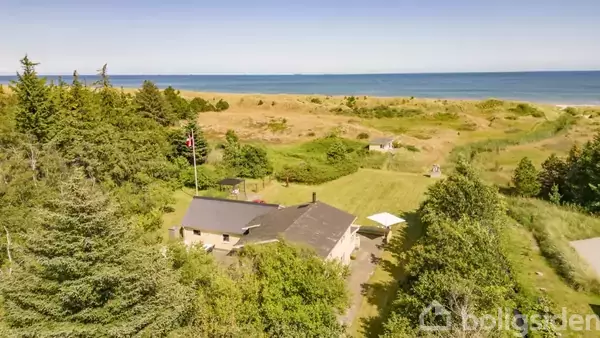 Hus omgivet af frodige træer og buske, beliggende nær en sandet kystlinje med udsigt over havet i baggrunden under en klar himmel.