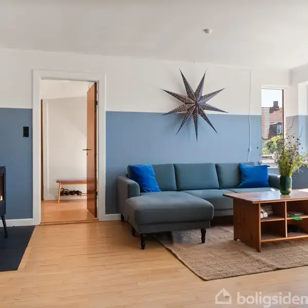 Sofa er placeret i en stue med blå vægge, flankeret af et sofabord og pyntet med puder. En brændeovn brænder i hjørnet. Store vinduer slipper lys ind.