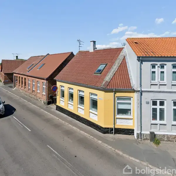 En hvid varebil kører på en vej flankeret af rækkehuse med røde tage; et gult hus står i forgrunden under en blå himmel med få skyer.