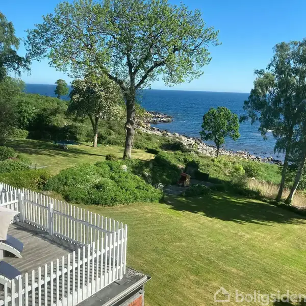 Terrasse med havemøbler set fra oven, omgivet af grøn plæne og spredte træer, der fører til en stenet kystlinje med udsigt over havet på en solskinsdag.