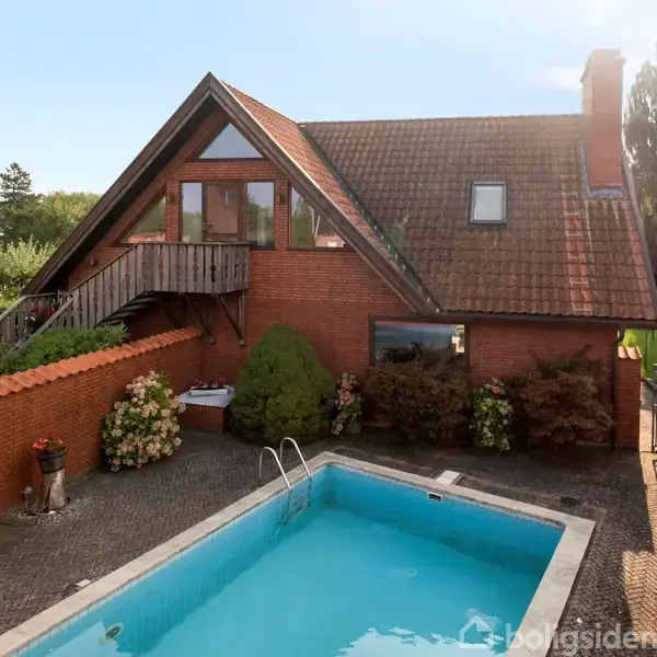 Et rødt murstenshus med trekantet gavl har en stor udendørs swimmingpool. Poolen er omgivet af fliser og grønne planter i en hyggelig baggård.