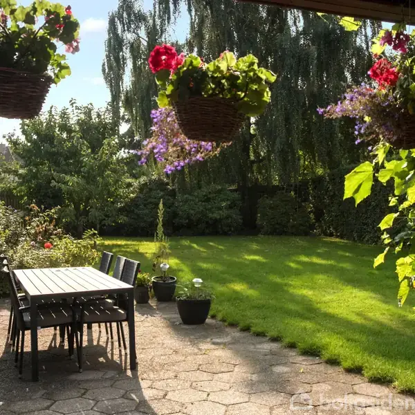 Hængende blomsterkurve pryder en terrasse, hvor et bord og stole står. Sollys belyser en grøn have i baggrunden med træer og buske.