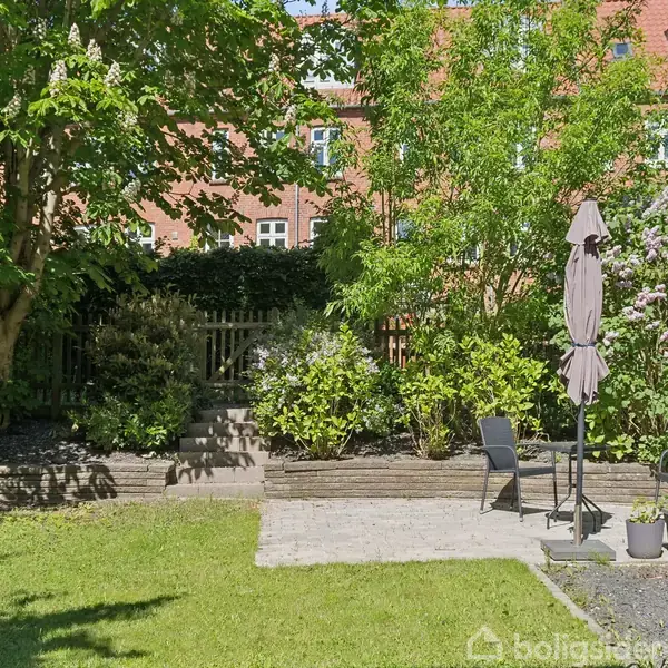 Have og terrasse med grøn græsplæne og stenterrasse. Parasol og stole står på terrassen. Omkring er der buske og træer, og en murstensbygning i baggrunden.