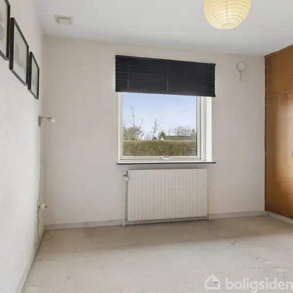 Et tomt værelse har træskabe til højre og en radiator under et vindue. Væggene er hvide med billeder hængende. Udenfor er træer og himmel synlige.