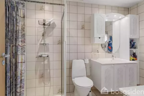 Badeværelse med brusekabine; væghængt bruser, forhæng til venstre. Toilet og vaskeskab med spejl og indbygget lys til højre. Flisevægge i neutrale toner, ren indretning.