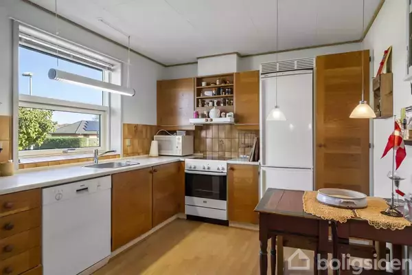 Køkken med træskabe, ovn og opvaskemaskine. Et lille bord med tallerkener står centralt. Udsigt til have gennem vindue. Lamper hænger ned fra loftet.