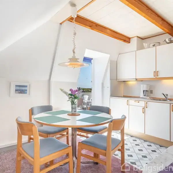 En rund spisebord med fire stole står foran et funktionelt køkken med hvide skabe. Over bordet hænger en lampe, og på bordet er en vase med blomster.