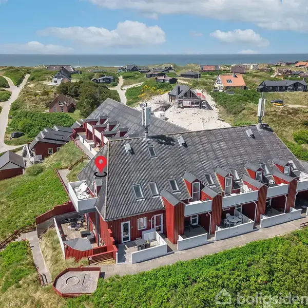 Bygning med flere røde murstenshuse har hvide altaner og ligger i et grønt klitlandskab. Baggrunden viser flere sommerhuse, sandstier og havet i horisonten.