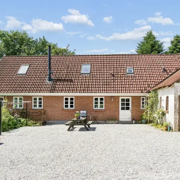 Et rødt murstenshus med rødt tegltag står stille i en solbelyst, grusbelagt gårdsplads, omgivet af grønne træer og en anden lille, hvidbygget bygning til højre.
