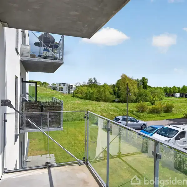 En balkon med glasrækværk ser ud over en parkeringsplads med biler og en grøn park med træer i baggrunden. To nabobalkoner ses også på bygningsfacaden.