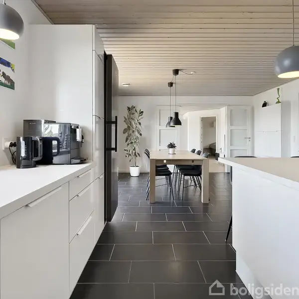 Et moderne køkken med hvide skabe indeholder en kaffemaskine og emhætte. Et langt spisebord med stole er i baggrunden. Rummet har grå fliser og hængende lamper.