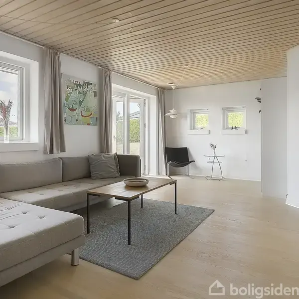 En moderne stue med en stor, grå sofa placeret ved vinduer. Et rektangulært sofabord står på et tæppe. Rummet er lyst med trægulv og loft.