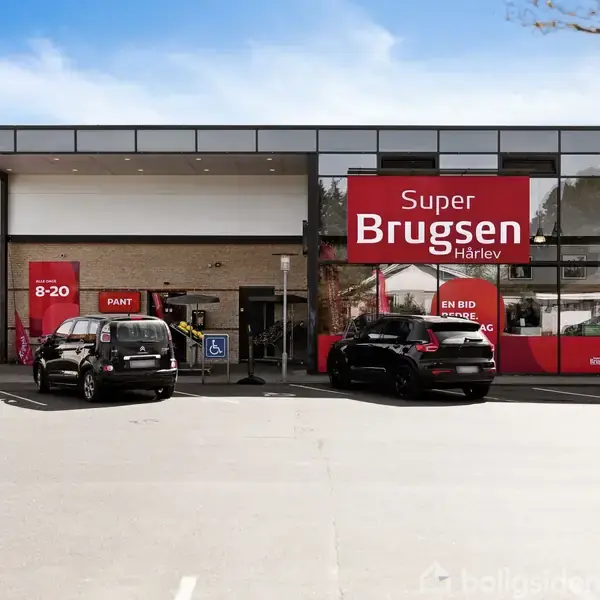 Supermarkedet SuperBrugsen i Hårlev står stille foran en parkeringsplads med biler. Tekst på skilt: "Super Brugsen Hårlev", "Pant", "En bid af Danmark", "8-20".