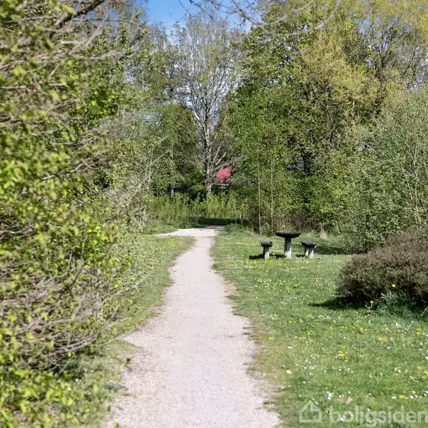 Sti snor sig gennem frodig park, omkranset af grønne buske og træer; en bænk og bord ses til højre. I baggrunden anes et rødt tag.