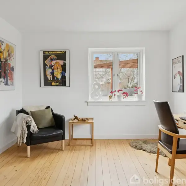 En stue med en sort lænestol og bord ved vinduet, skrifter med "Le Mans" på plakaten på væggen. Lyse trægulve, hvidmalede vægge, bøger på hylden.